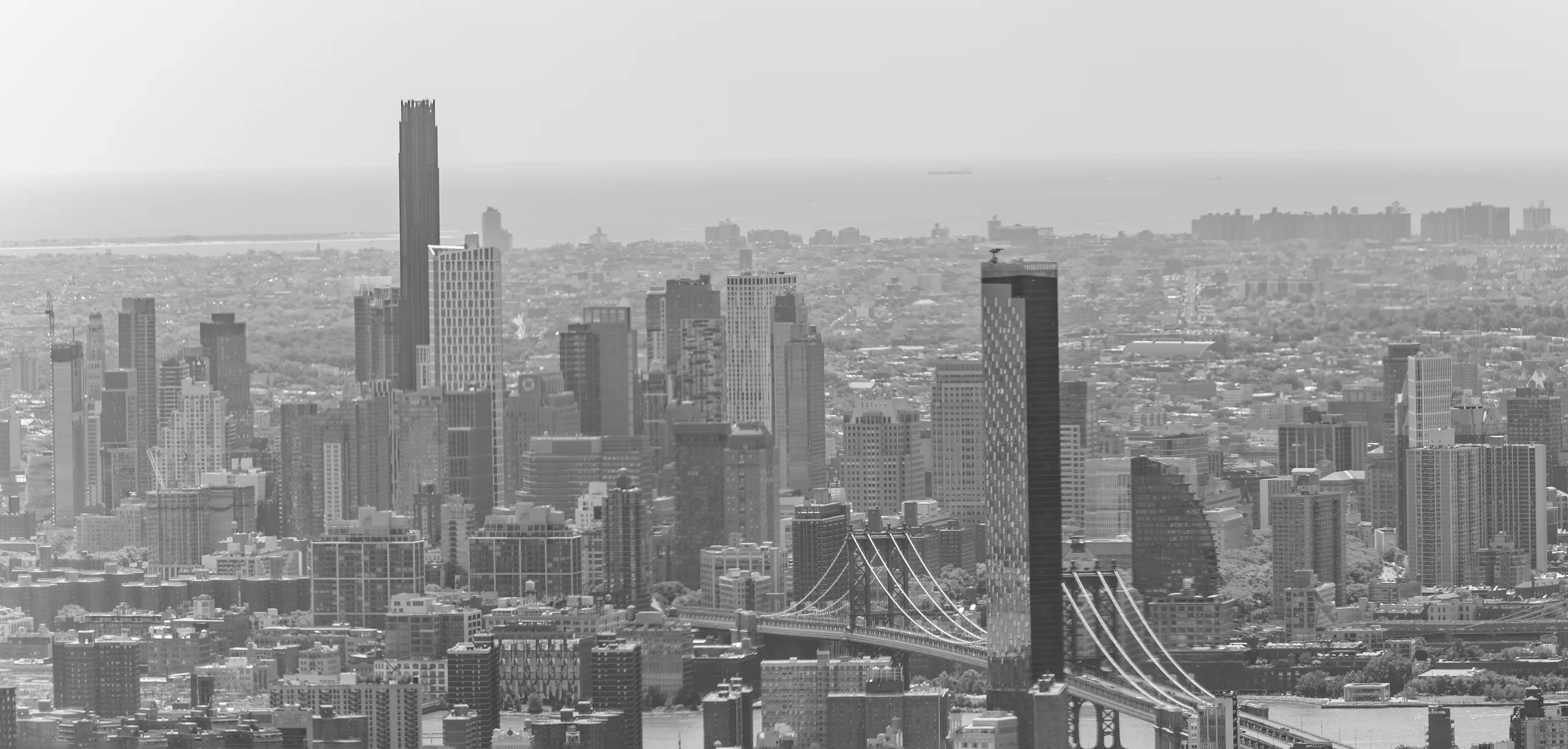 Black and white view south across Manhattan rooftops toward the Financial District, with a suspension bridge crossing the East River at the right.