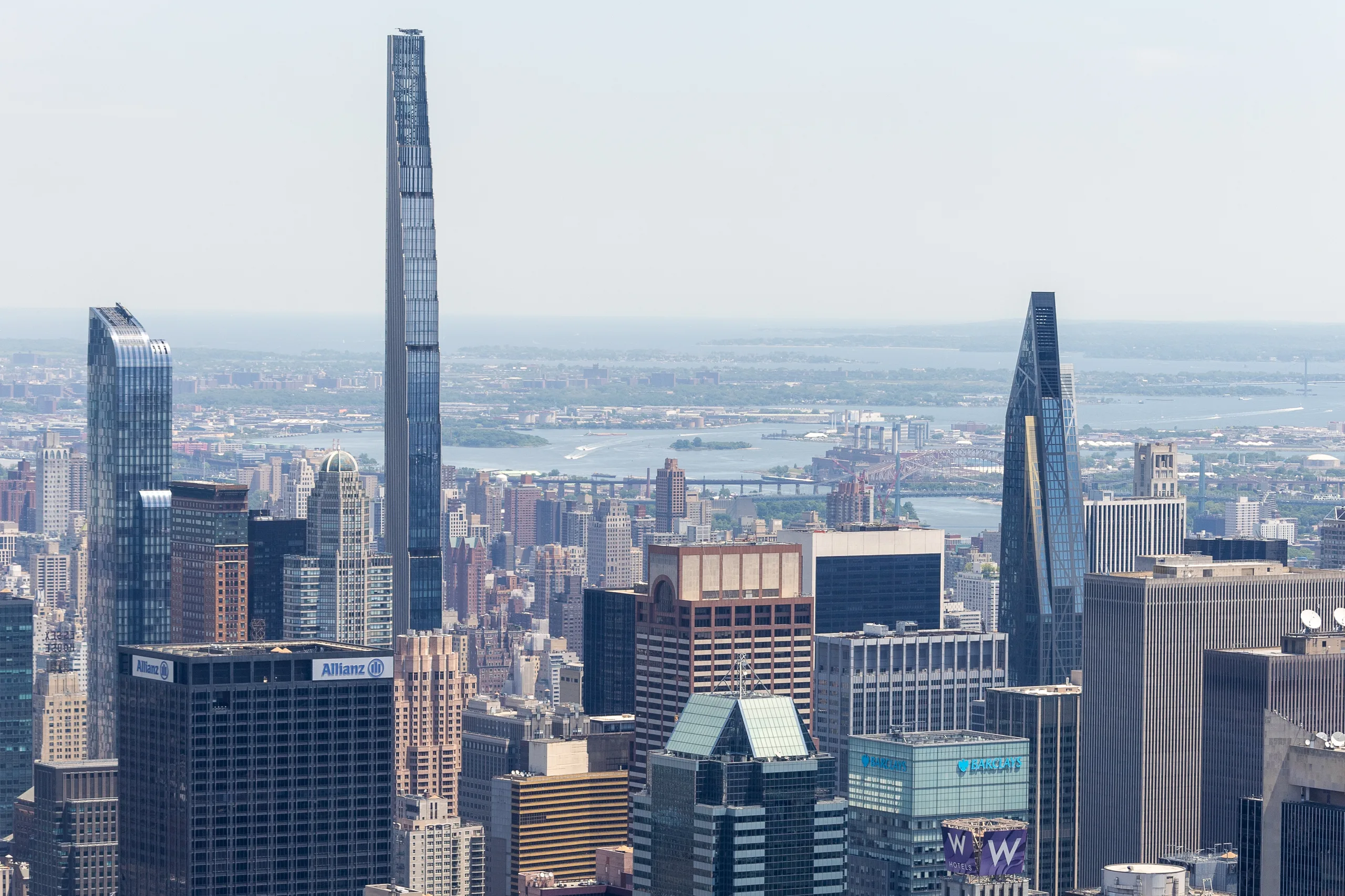 Midtown Manhattan rooftops in daylight with the slender supertall towers of Billionaires' Row rising above them and the harbour on the horizon.