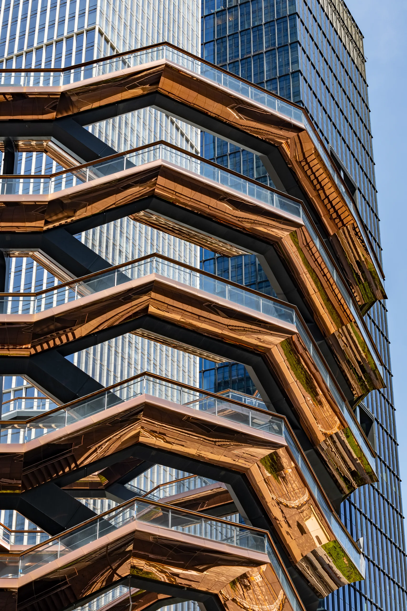 Close view of the copper-clad interlocking staircases of Vessel at Hudson Yards, with a glass tower rising behind it.