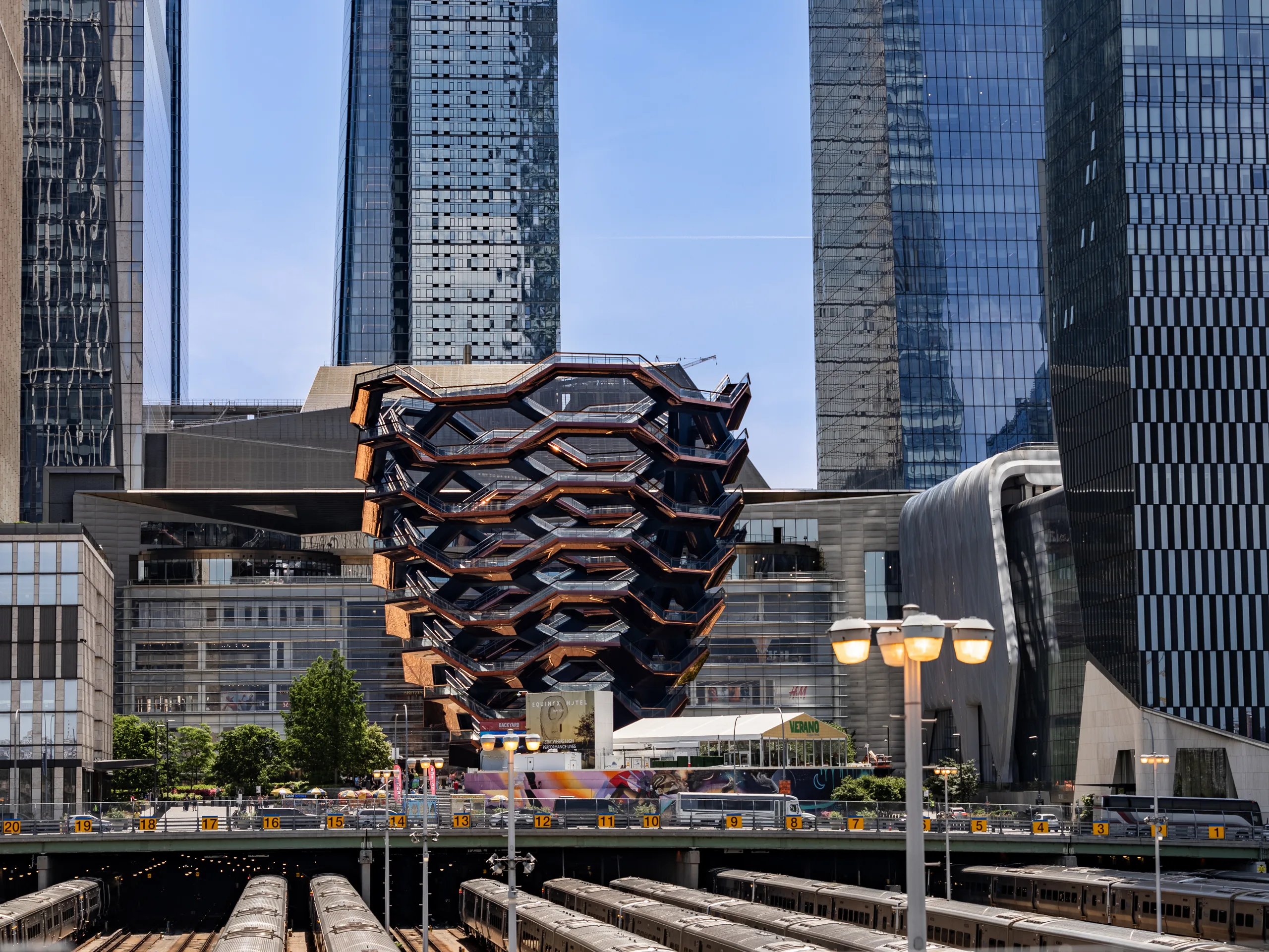 Vessel at Hudson Yards seen from above the West Side rail yards, framed by mirrored glass towers on both sides.